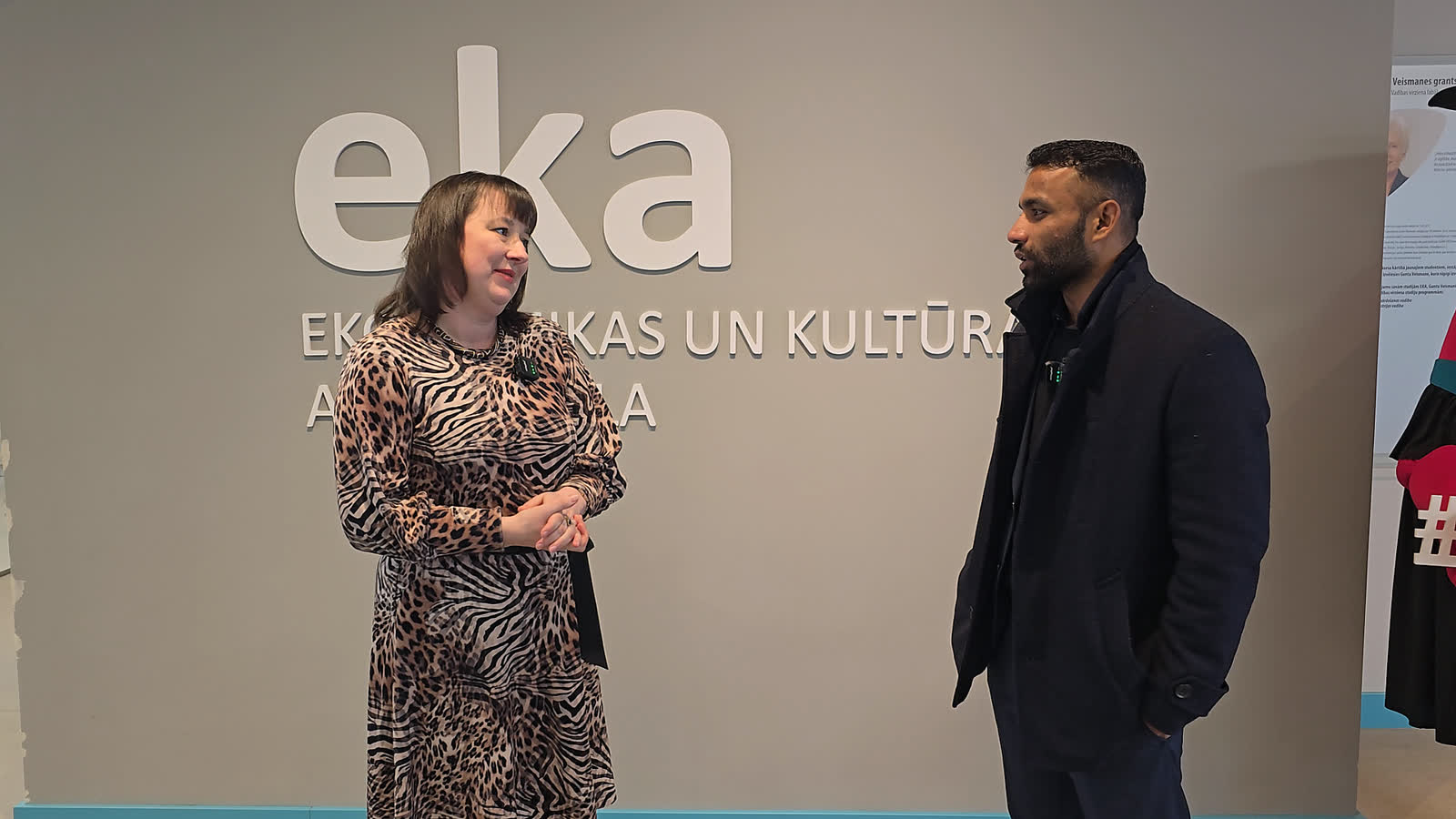 An EKA representative and our CEO standing in conversation in front of the lettered wall reading “eka — EKONOMIKAS UN KULTŪRAS AUGSTSKOLA”