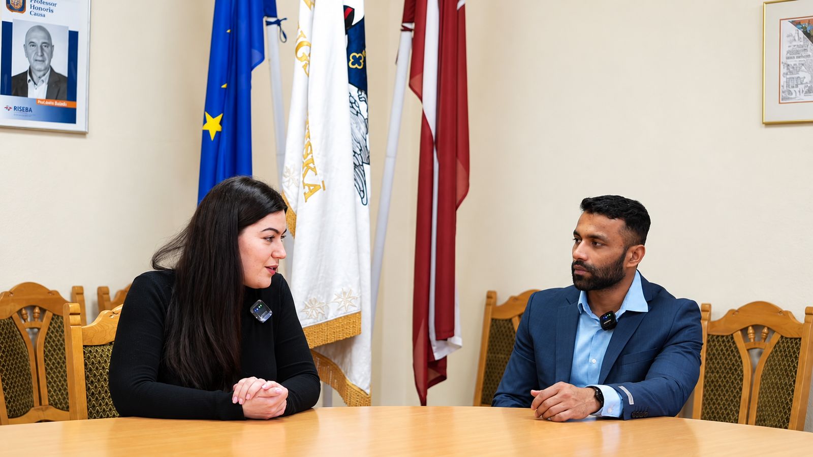 A RISEBA representative and our colleague in conversation at a meeting table, beneath the EU, RISEBA and Latvian flags and RISEBA's “Professor Honoris Causa” portraits
