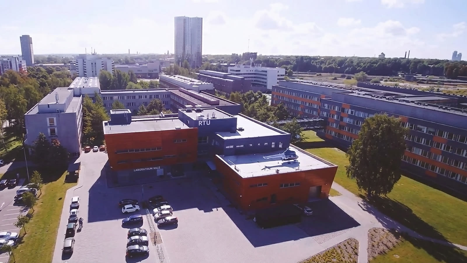 RTU's campus on Ķīpsala from the air: a red and blue building with “RTU” on it, the car park in front and Riga's towers behind