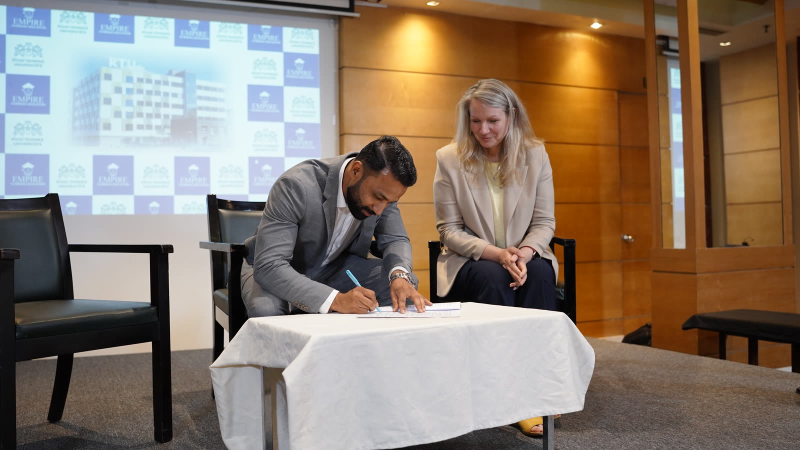 Our colleague signing a document at a small table beside a guest from RTU, in front of a screen showing our logo and an RTU building