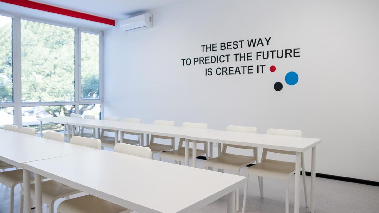 A bright classroom at Turība with white tables and chairs, and the words “The best way to predict the future is create it” on the wall